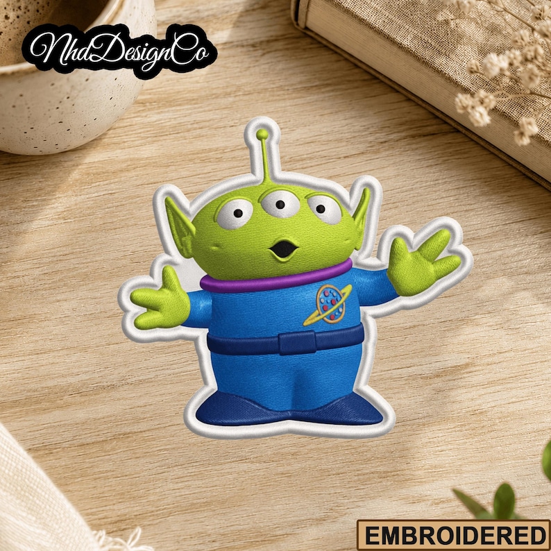 May include: Embroidered patch featuring a green, three-eyed alien character in a blue uniform with a purple collar. The patch has a white border and the text "EMBROIDERED" is at the bottom. The patch is on a wooden surface.