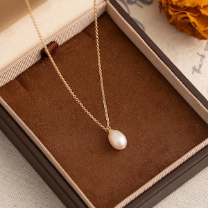 May include: A delicate gold chain necklace with a single, pear-shaped pearl pendant. The necklace is displayed in a brown velvet jewelry box.