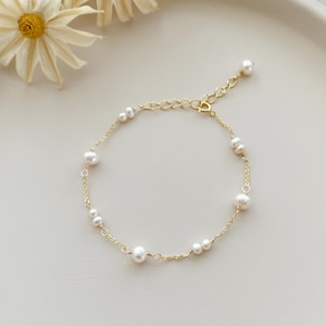 Könnte beinhalten: Ein zartes Armband aus goldener Kette mit kleinen weißen Perlen. Das Armband hat einen Karabinerverschluss.