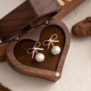 May include: A pair of pearl earrings with gold bow accents are displayed in a heart-shaped wooden box.