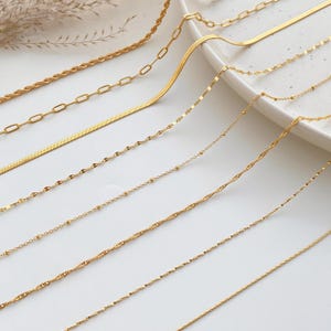 Puede incluir: Una colección de collares dorados en varios estilos, incluyendo diseños de cuerda, eslabones y cadena de serpiente. Los collares se exhiben sobre una superficie blanca, mostrando sus diferentes texturas y diseños. Las joyas se presentan en un entorno bien iluminado.