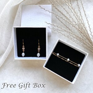 May include: Two white gift boxes with black interiors. One box contains a pair of pearl and crystal earrings. The other box contains a delicate gold chain necklace with three pearls.