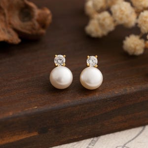 May include: A pair of pearl earrings with a small clear rhinestone on top of each pearl.