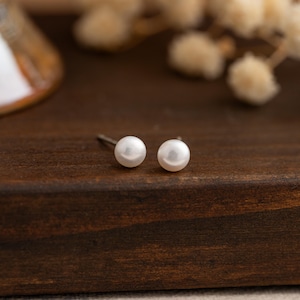 May include: A pair of small, round, white pearl stud earrings. The earrings are set on silver posts and sit on a dark brown wooden surface. The pearls have a smooth, lustrous finish. The background includes blurred white floral accents.