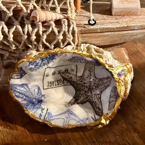 May include: A white seashell with gold trim, decorated with a blue and white design featuring a starfish and the words "C POSTAGE".