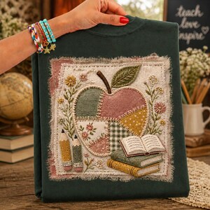 Op de afbeelding: Een donkergroen sweatshirt met een vierkante patch met een patchwork appelontwerp, twee potloden en een stapel van twee open boeken. De appel is samengesteld uit verschillende stofpatronen, waaronder bloemen, ruitjes en effen kleuren met stiksels.