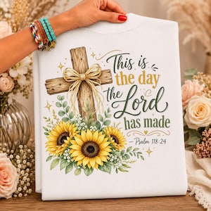 May include: A white t-shirt featuring a graphic of a wooden cross adorned with a gold ribbon bow and yellow sunflowers. The text reads, "This is the day the Lord has made - Psalm 118:24" in a mix of cursive and serif fonts with small gold stars.