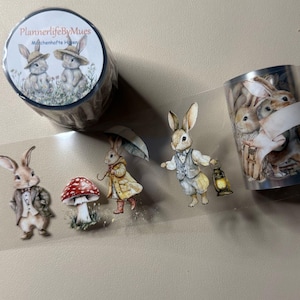 Könnte beinhalten: Eine Rolle transparentes Washi-Tape mit einem sich wiederholenden Muster von Hasen, die Hüte tragen und Regenschirme, Pilze und Laternen halten. Das Band ist auf einer Spule verpackt mit einem Etikett, auf dem "PlannerlifeByMues" und "Mädchenhafte Hüpfer" steht.