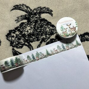 Op de afbeelding: Een rol washi tape met een witte achtergrond en een patroon van groene bomen en bruine herten. De tape ligt op een wit oppervlak en de rol zit in een doorzichtige plastic verpakking met de tekst "PlannerlifebyMuses" en "Woodland Vol.2" erop gedrukt.