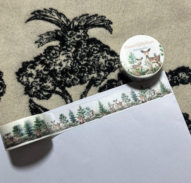 Op de afbeelding: Een rol washi tape met een witte achtergrond en een patroon van groene bomen en bruine herten. De tape ligt op een wit oppervlak en de rol zit in een doorzichtige plastic verpakking met de tekst "PlannerlifebyMuses" en "Woodland Vol.2" erop gedrukt.