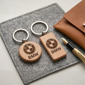 May include: Two wooden BMW keychains, one round and one rectangular, with silver key rings. The BMW logo and text are engraved on the light-colored wood. They rest on a gray felt surface, with a leather pouch and pen nearby.