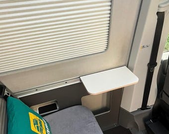 May include: A white folding table is mounted to the wall inside a van. The table is attached to a gray wall panel with a silver trim. The table is folded up and is not in use.