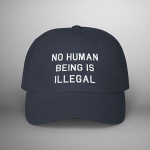 May include: Navy blue baseball cap with the white text "NO HUMAN BEING IS ILLEGAL" across the front. The cap has a curved brim and a rounded crown. A simple and direct statement.