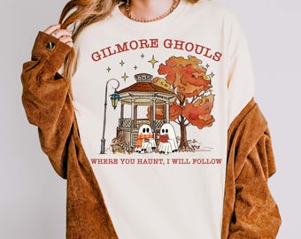 May include: Cream-colored sweatshirt with a graphic featuring the text "GILMORE GHOULS" above an illustration of a gazebo, autumn tree, and two ghost characters. The text "WHERE YOU HAUNT, I WILL FOLLOW" is below the graphic.