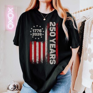 Pode incluir: Camiseta preta com um design desgastado da bandeira americana com listras vermelhas e brancas e estrelas. A camisa inclui o texto "1776-2026" e "250 YEARS" em branco. Uma pe&ccedil;a de roupa casual e patri&oacute;tica.