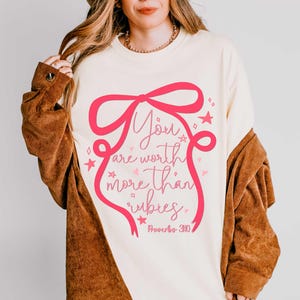 May include: Cream-colored sweatshirt with a pink bow and the text "You are worth more than rubies" in a script font. The design includes stars and hearts. The sweatshirt is part of a clothing collection.
