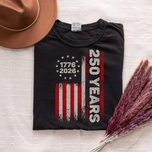 Pode incluir: Camiseta preta com um design desgastado da bandeira americana. A bandeira inclui os anos de 1776 e 2026, e o texto "250 YEARS" em branco. Um chap&eacute;u marrom e um feixe de grama seca, avermelhada-p&uacute;rpura, tamb&eacute;m est&atilde;o na imagem.