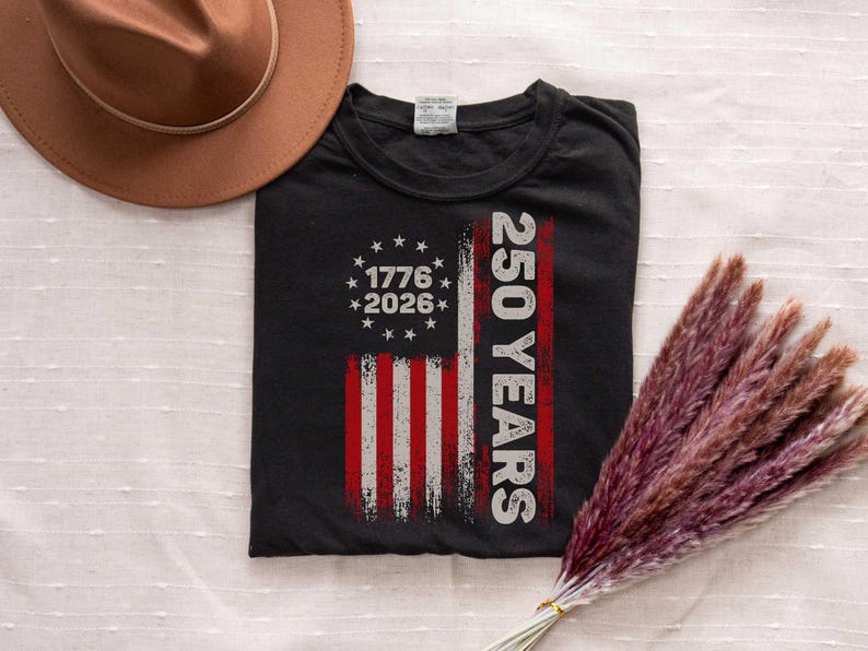 Pode incluir: Camiseta preta com um design desgastado da bandeira americana. A bandeira inclui os anos de 1776 e 2026, e o texto "250 YEARS" em branco. Um chap&eacute;u marrom e um feixe de grama seca, avermelhada-p&uacute;rpura, tamb&eacute;m est&atilde;o na imagem.