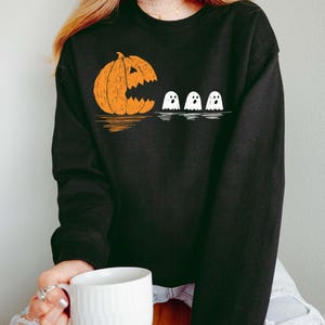Puede incluir: Sudadera negra con un diseño de Halloween. El gráfico muestra una calabaza naranja con una cara tallada 'comiendo' tres figuras de fantasmas blancos. Una prenda casual y cómoda para la temporada de miedo.