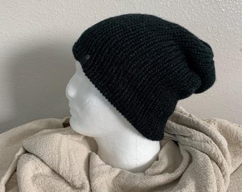 May include: A dark green knit beanie with a small, circular tag on the side. The hat is displayed on a white mannequin head, with a neutral-colored fabric in the background.