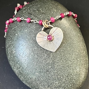 May include: A silver heart pendant necklace with a small, round, red gemstone in the center. The pendant has a sunburst design. The necklace features a chain of small, pink beads. The necklace is displayed on a gray stone.