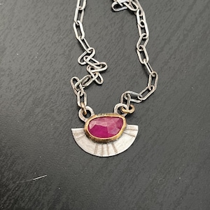 May include: A silver chain necklace with a pendant featuring a pink gemstone set in a silver and gold setting. The pendant is shaped like a crescent moon with a textured surface.