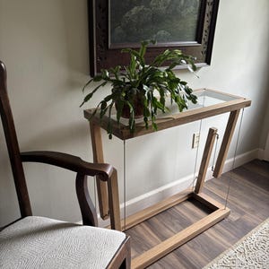May include: A wooden tension table featuring a rectangular glass top supported by a floating frame structure held together by thin vertical wires, with a green potted plant placed on the surface and a wooden chair with a light fabric seat nearby.