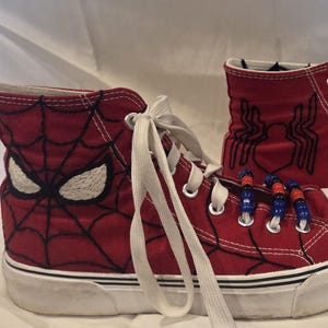May include: Red high-top sneakers featuring Spider-Man designs. One shoe displays the character's eyes and web pattern, while the other showcases the spider emblem. White laces and soles complete the look. The shoes have a white toe cap and are decorated with blue and red beads.