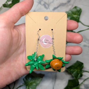 May include: A pair of dangle earrings featuring a green succulent and an orange fruit charm. The earrings are on a silver chain and have silver hooks.