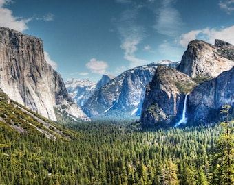 May include: A panoramic view of a valley with towering rock formations and a waterfall cascading down the side of a cliff. Lush green trees cover the valley floor, creating a vibrant contrast against the gray rock faces.