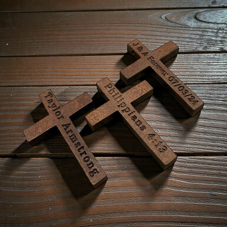 May include: Three wooden crosses with engraved text. One cross reads "Taylor Armstrong", another "Philippians 4:13", and the third "J & A Forever 07/03/24". The crosses are a medium brown color and rest on a wooden surface.