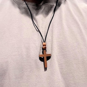 May include: A wooden cross pendant necklace with a dark cord. The cross is a medium brown color and is suspended from a black cord. Two wooden beads are above the cross. The necklace is worn over a light gray t-shirt.