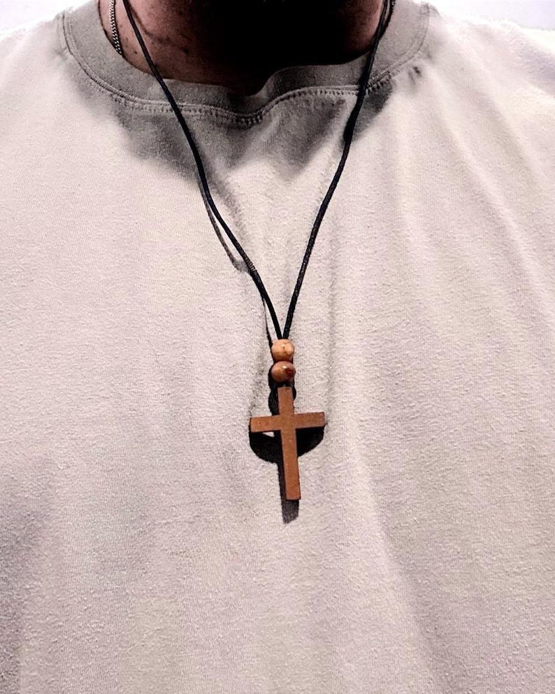 May include: A wooden cross pendant necklace with a dark cord. The cross is a medium brown color and is suspended from a black cord. Two wooden beads are above the cross. The necklace is worn over a light gray t-shirt.