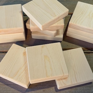 May include: Several unfinished square wooden coasters are arranged on a wooden surface. The coasters are light-colored with visible wood grain. Some are stacked, while others are spread out, showcasing their simple, natural design.