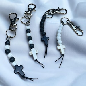 May include: Four keychains with religious themes. Each keychain features a silver clasp, a beaded strand of white and black beads, and a cross pendant. The crosses are either white or black. The keychains are displayed on a white fabric background.