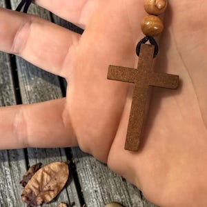 May include: A wooden cross pendant on a black cord necklace. The cross is a medium brown color and has a simple, classic design. Two wooden beads are above the cross. The necklace is held in a hand.
