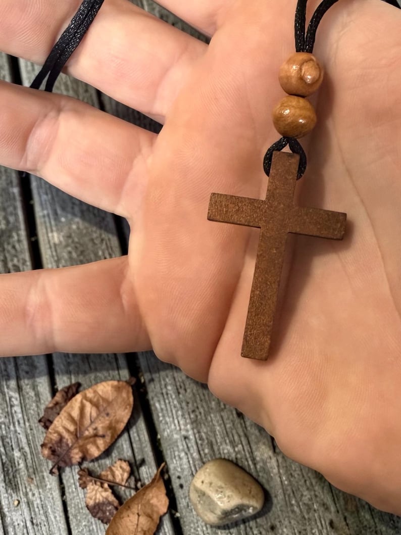 May include: A wooden cross pendant on a black cord necklace. The cross is a medium brown color and has a simple, classic design. Two wooden beads are above the cross. The necklace is held in a hand.