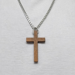 May include: A wooden cross pendant hangs from a silver chain. The cross is a warm brown color and has a simple, classic design. The chain is a curb style and is worn against a white shirt.