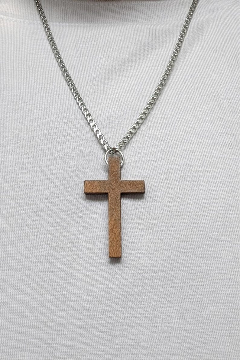 May include: A wooden cross pendant hangs from a silver chain. The cross is a warm brown color and has a simple, classic design. The chain is a curb style and is worn against a white shirt.