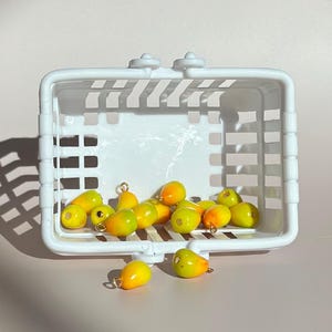 May include: A white plastic basket filled with yellow and orange pear-shaped charms. The charms are scattered inside and outside the basket. The background is a light pink surface with shadows.