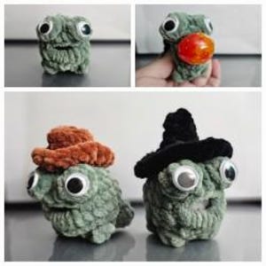 May include: A green plush toy with large eyes and a textured surface. The toy is wearing a brown felt hat.