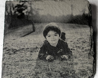 Puede incluir: Un posavasos de pizarra grabado en blanco y negro con un niño tumbado en la hierba. La imagen es un retrato de un niño pequeño, con un fondo borroso. El posavasos tiene un borde natural y rugoso.