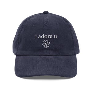 May include: Navy blue corduroy baseball cap with the phrase "i adore u" embroidered in white, along with a small flower graphic. The cap has a curved brim and an adjustable strap at the back.