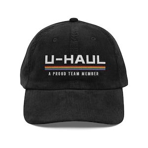 Op de afbeelding: Zwarte corduroy baseballcap met het U-HAUL-logo in witte blokletters boven een regenboogstreep en de tekst "A PROUD TEAM MEMBER". De cap heeft een gebogen klep en een knoop aan de bovenkant.