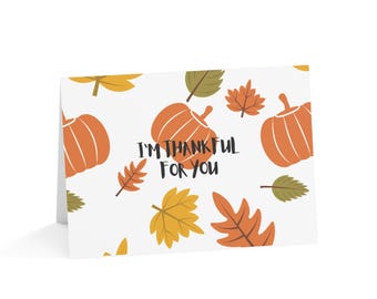May include: A white greeting card with a fall theme. The card features orange pumpkins and autumn leaves in shades of orange, yellow, and green. The text "I'M THANKFUL FOR YOU" is printed in black.