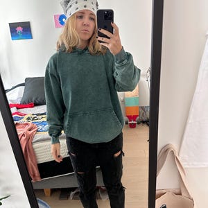 K&ouml;nnte beinhalten: Eine Person tr&auml;gt ein gr&uuml;nes Kapuzenpullover, eine schwarze zerrissene Jeans und wei&szlig;e Turnschuhe. Sie tr&auml;gt ein wei&szlig;es Bandana mit einem schwarzen Paisley-Muster auf dem Kopf. Die Person macht ein Selfie in einem Spiegel.