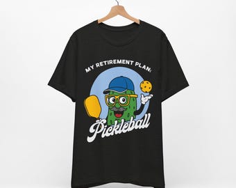 May include: Black t-shirt with a cartoon pickle character wearing glasses and a cap, holding a pickleball paddle and ball. The text says "MY RETIREMENT PLAN: Pickleball".