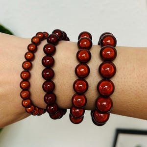 Könnte beinhalten: Vier elastische Armbänder mit glatten, runden, dunkelroten Perlen in glänzender Optik. Die Armbänder unterscheiden sich in der Perlengröße, wobei die beiden linken kleinere Perlen und die beiden rechten größere, einheitliche Kugeln aufweisen.