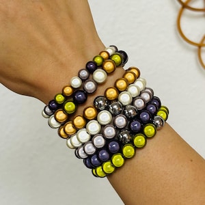 Könnte beinhalten: Ein Stapel aus mehreren Perlenarmbändern mit runden Perlen in verschiedenen Farben, darunter Limettengrün, Metallic-Gold, Dunkelviolett, Weiß und Dunkelgrau. Die Armbänder sind in einem geschichteten Muster am Handgelenk angeordnet und zeigen eine Mischung aus glänzenden und metallischen Oberflächen auf den kugelförmigen Perlen.