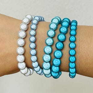 Könnte beinhalten: Ein Set aus vier elastischen Armbändern mit runden Perlen in einem Farbverlauf. Das erste Armband ist weiß, das zweite hellblau, das dritte mittel-türkis und das vierte leuchtend cyanblau, alle mit einer glänzenden Oberfläche versehen.